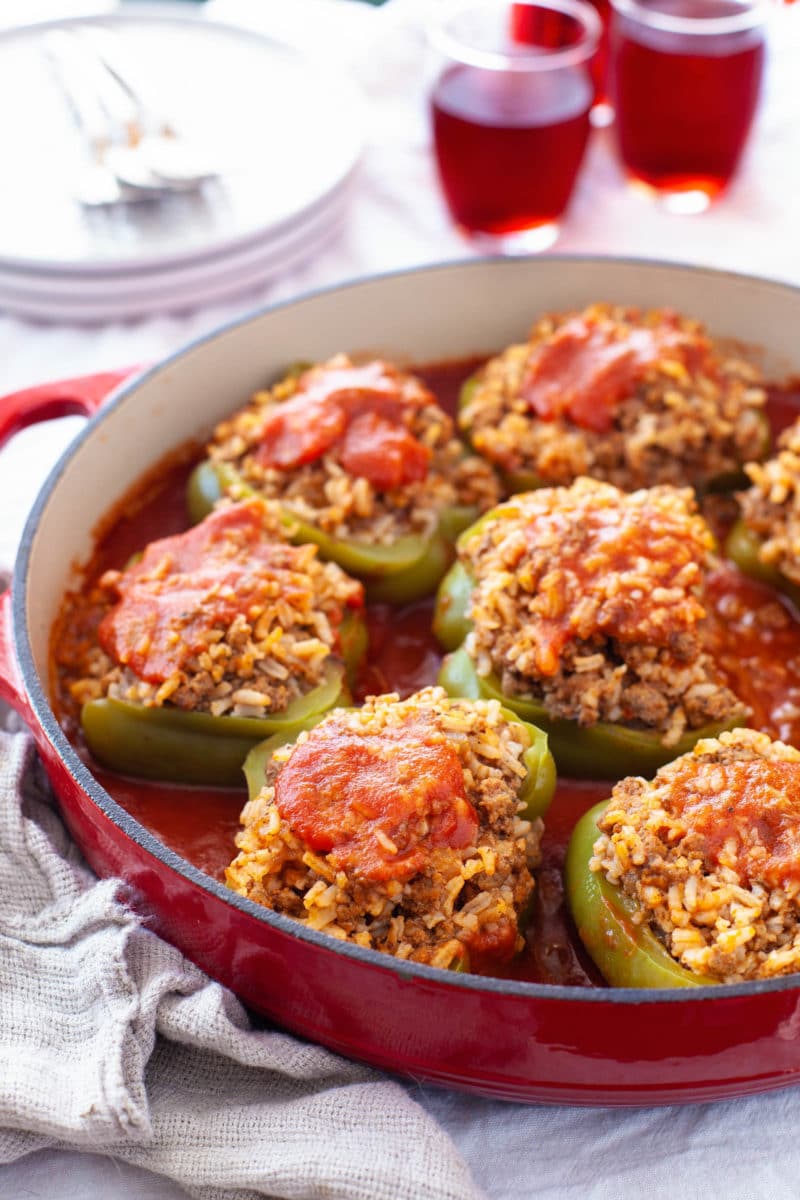 A shallow red Dutch oven filled with beef and rice stuffed bell peppers in a red sauce with a stack of white plates, forks, and glasses of red wine in the background