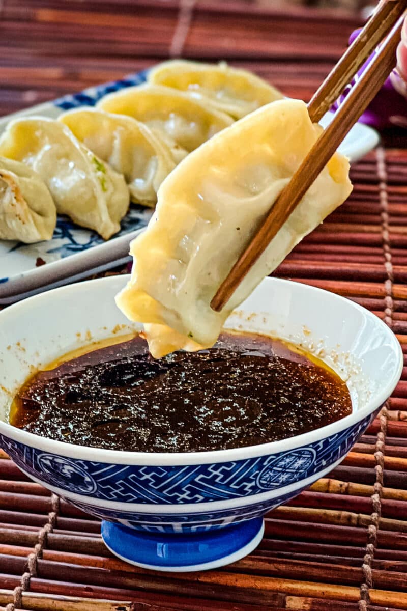 Chopsticks holding a cooked gyoza and dipping it into a blue bowl filled with potsticker sauce.