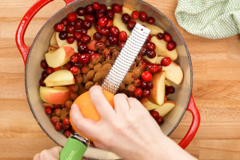 zesting an orange with a microplane grater over a pot of cranberry applesauce ingredients