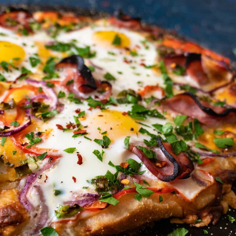 Breakfast pizza topped with crispy ham, sunny side up eggs, red bell pepper, red onion, green onion, parsley, and red pepper flakes