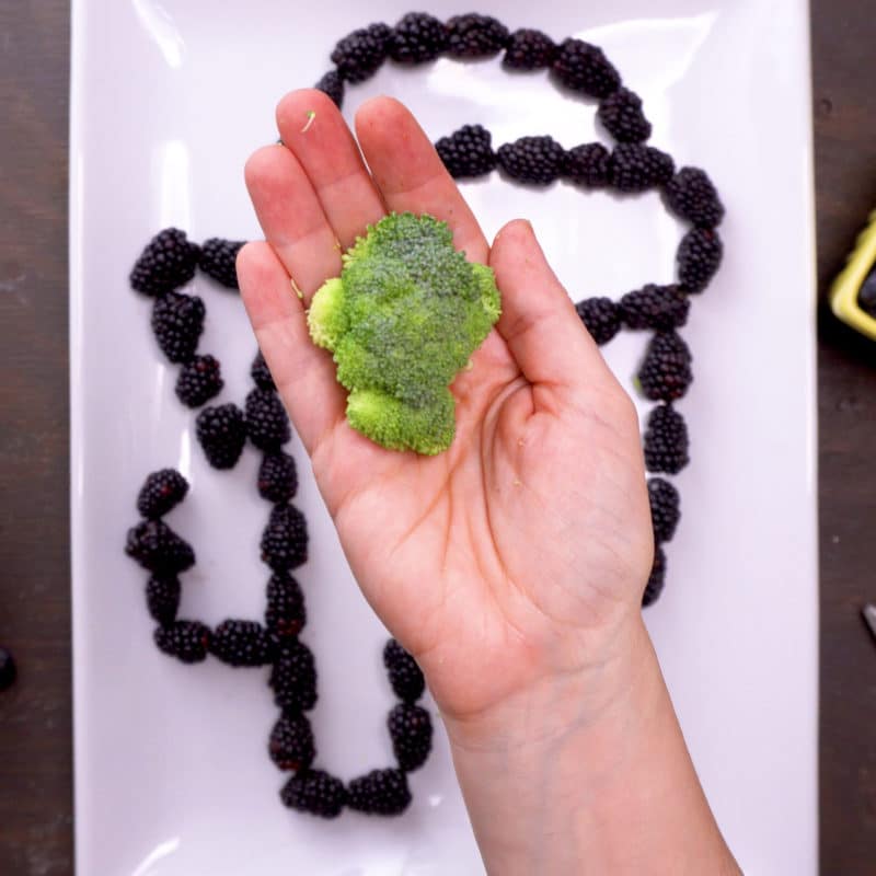 holding broccoli sitting flat in hand above an outline of Among Us crewmate made of blackberries