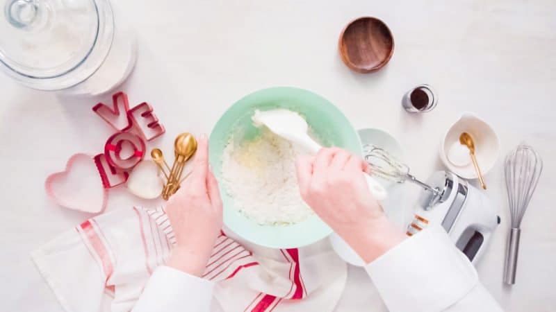 Hands mixing sugar cookie ingredients in a green bowl with heart cookie cutters on the counter