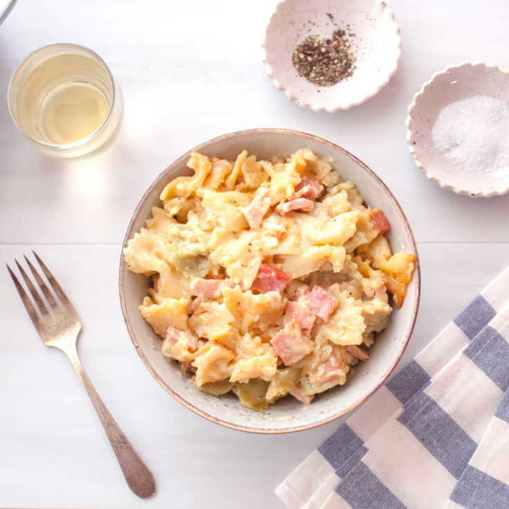 Bowtie pasta and ham in a creamy cheese sauce in a grey bowl with a blue and white striped napkin and glass of white wine