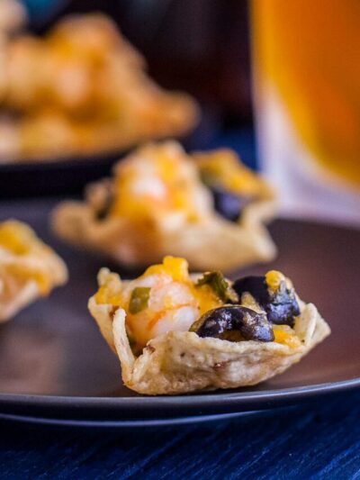 Close up of a shrimp, cheese, and olive filled tortilla scoop with another shrimp nacho and a beer in the background.