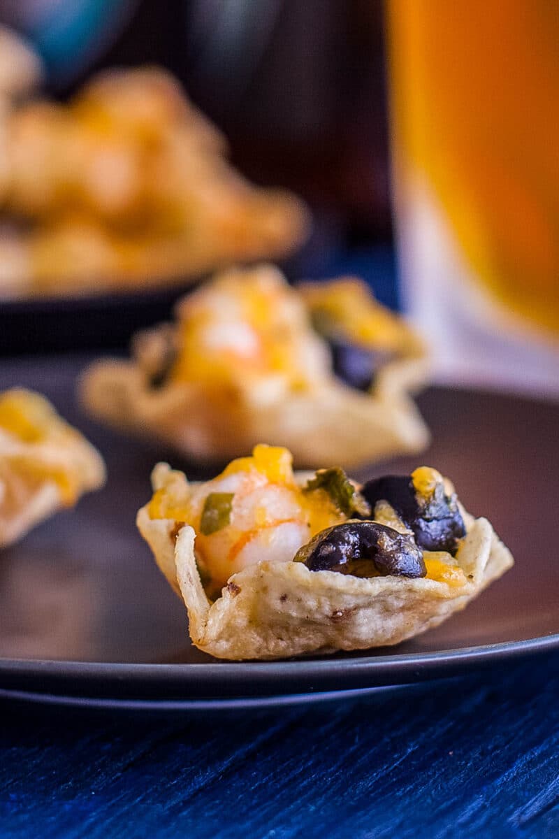 Close up of a shrimp, cheese, and olive filled tortilla scoop with another shrimp nacho and a beer in the background.