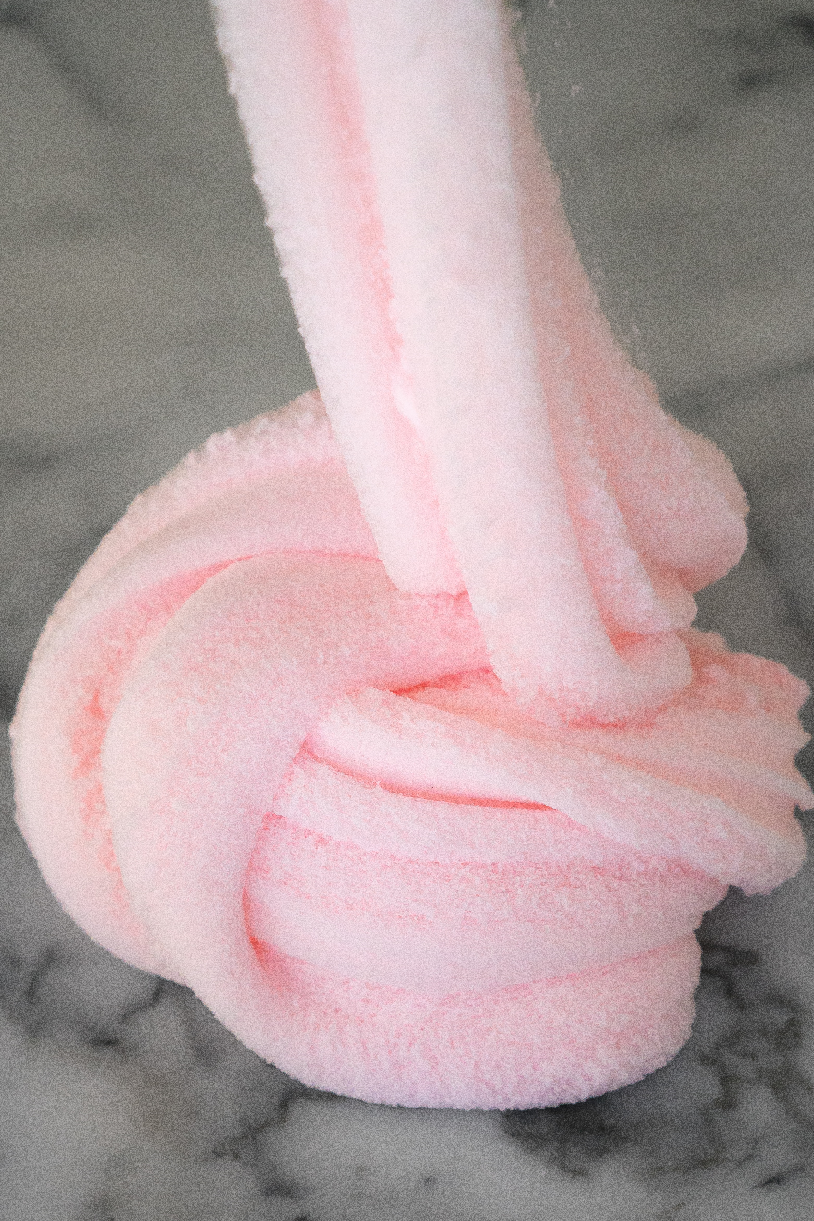 Fluffy pale pink cotton candy cloud slime drizzles down into a swirly pile on a white and grey marble counter.