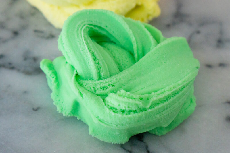 A swirly pile of green cloud creme slime sits on a white and grey marble board. The slime looks buttery and creamy, like a thick buttercream frosting.