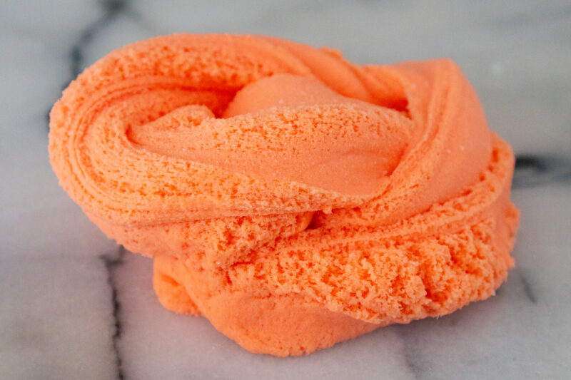 An orange cloud dough slime is swirled into a pile on a white and gray marble board. The edges of the ribbons of slime are filled with tiny little fissures like a chilled compacted sugar cookie dough.
