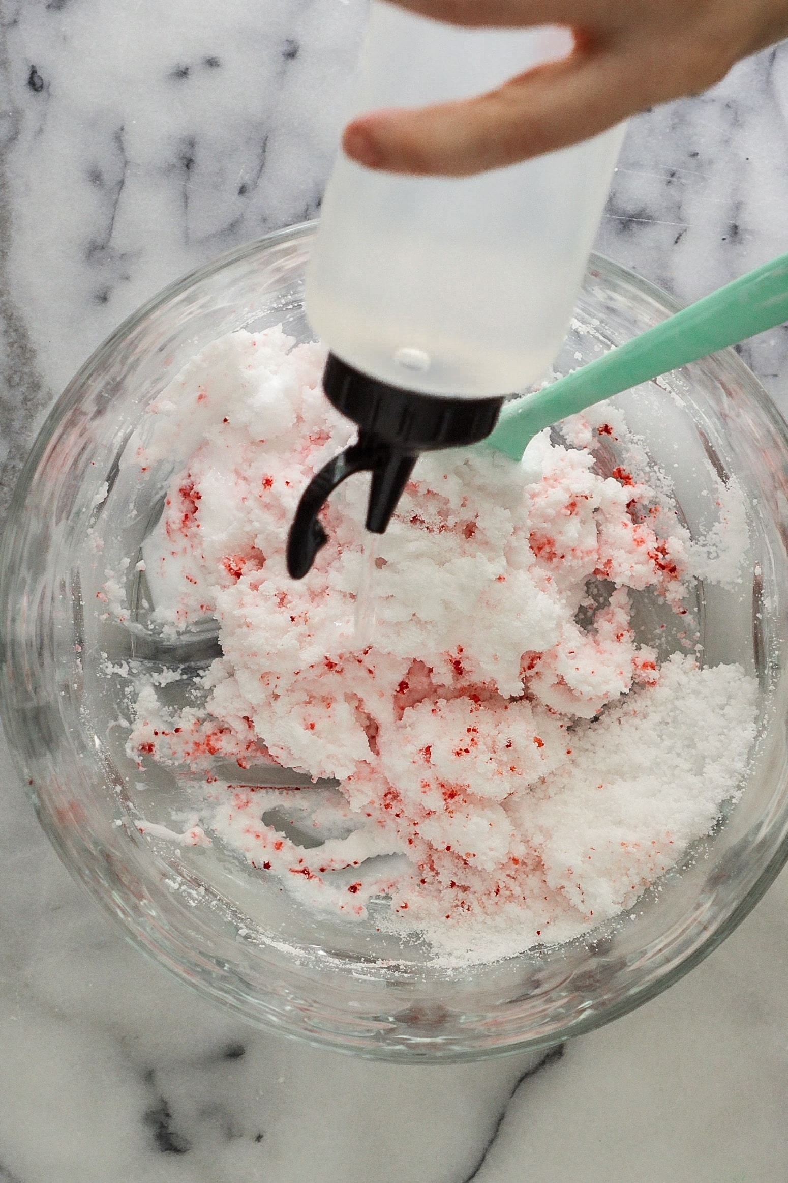 A clear condiment bottle with a black lid is filled with clear liquid slime activator and being squirted into a glass bowl of chunky slightly mixed slime ingredients.