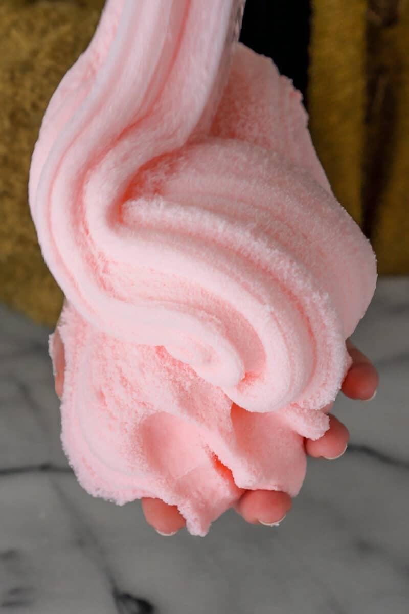 A woman's hand holds a large pile of incredibly fluffy pink cloud slime drizzling down in a swirl and looking just like cotton candy.