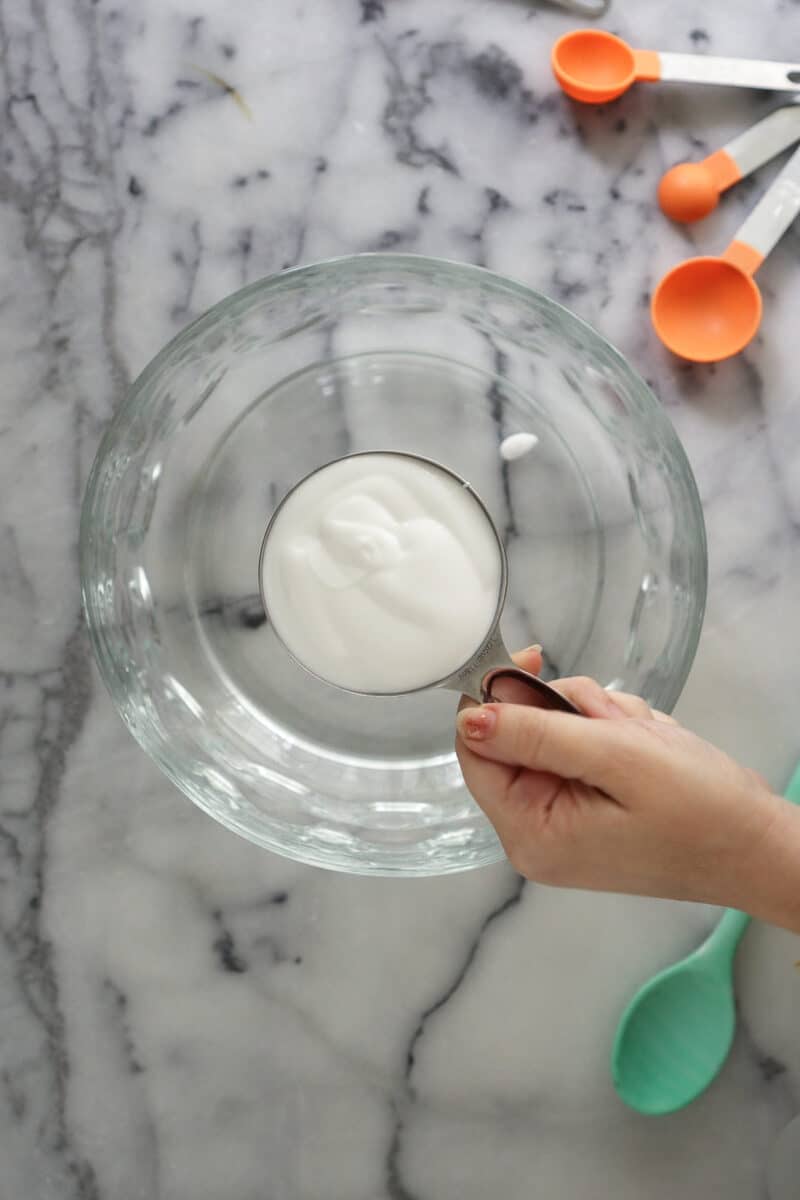 A woman's hand holds a silver measuring cup filled with white glue above an empty clear glass bowl. The bowl rests on a white and grey marble counter along with some orange measuring spoons and a mint green silicone spoon.
