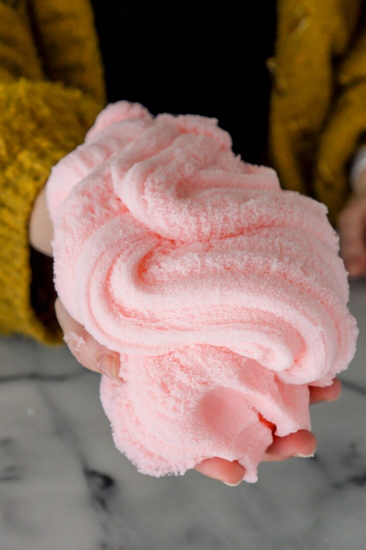 Close up of a large fluffy pile of pink cloud slime swirled in a woman's hand. She is wearing a black shirt and dark mustard yellow sweater and holding the slime above a white marble counter.