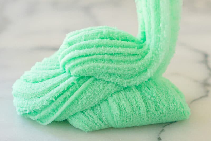 An incredibly fluffy ribbon of sea foam green cloud slime drizzles down into a swirly pile on a white marble counter. The texture of the slime looks like spun cotton candy.