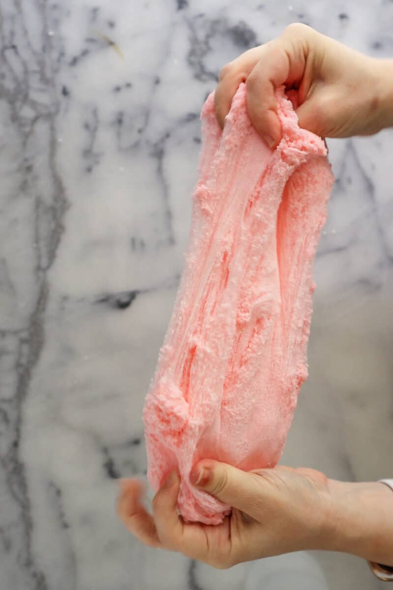 A woman's hands hold a pink cloud slime over a white and grey marble counter, stretching it out.